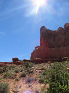 Majestic rock mountain in a National Park in Utah, with the sun shining brightly at the top. In the foreground, there is typical desert vegetation such as bushes and grasses growing on the dry ground.