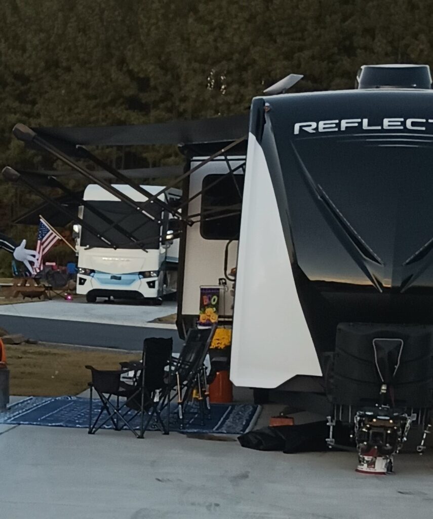 RVs parked in a luxury RV park. There is a travel trailer in the foreground and a motorhome to the left behind it on a separate RV site. There are woods behind the motorhome. 