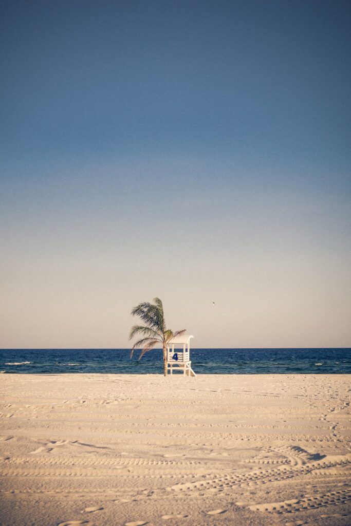 Lifeguard Chair situated on white sand at Gulf Shores, Alabama. The chair is elevated above the sand and has a tall backrest with a palm tree behind it. 